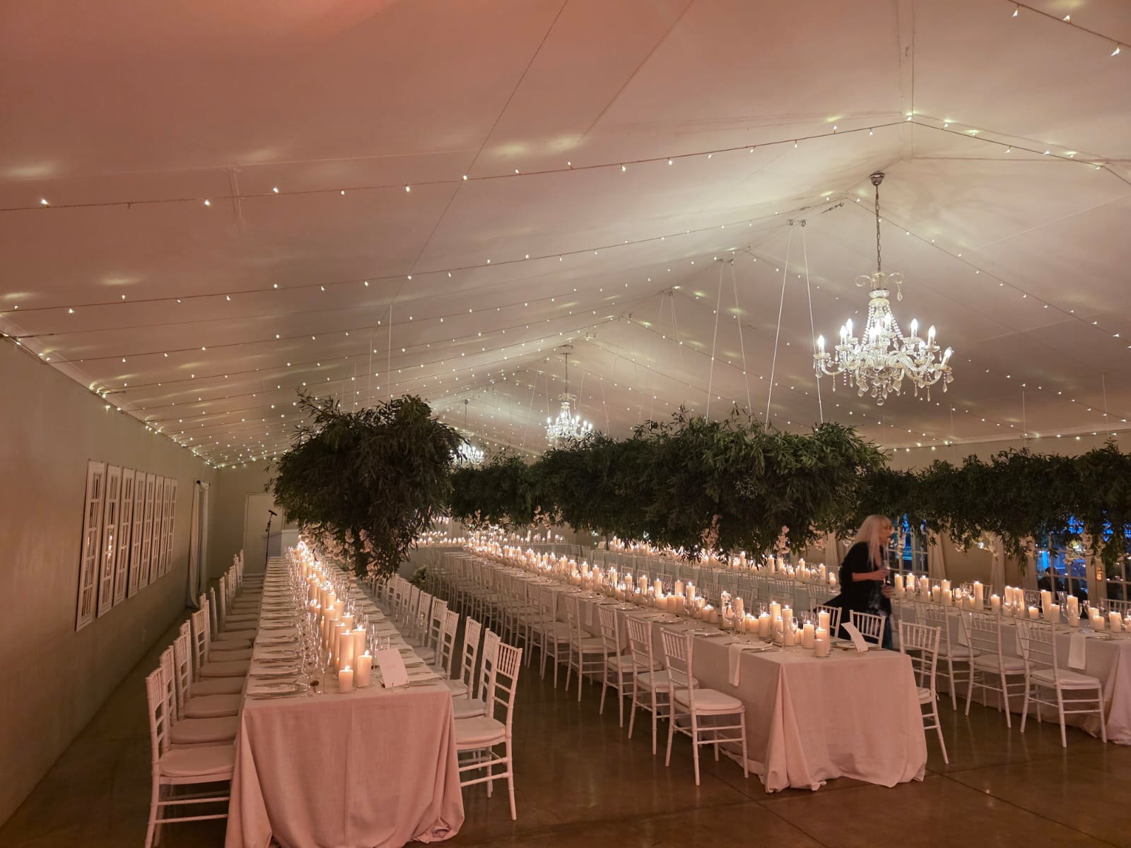 Long banquet table setup at Providence Country Weddings with blush pink tablecloths, rows of pillar candles down center, hanging greenery installations overhead, fairy lights and chandeliers creating romantic candlelit wedding dinner atmosphere