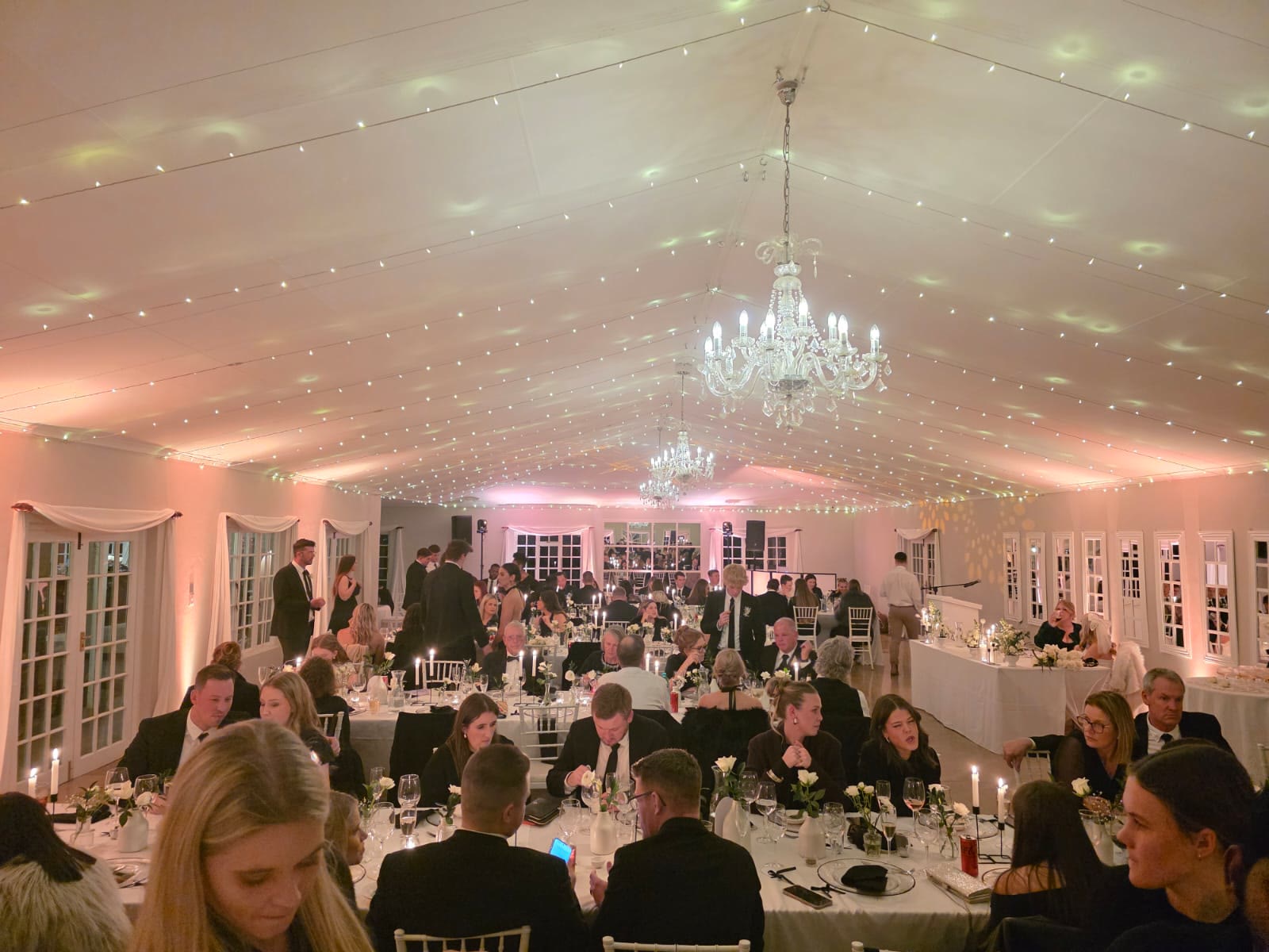 Wedding guests seated for dinner at Providence Country Weddings reception with romantic pink uplighting throughout hall, crystal chandeliers, fairy lights on ceiling, round tables with white Tiffany chairs creating warm elegant ambiance