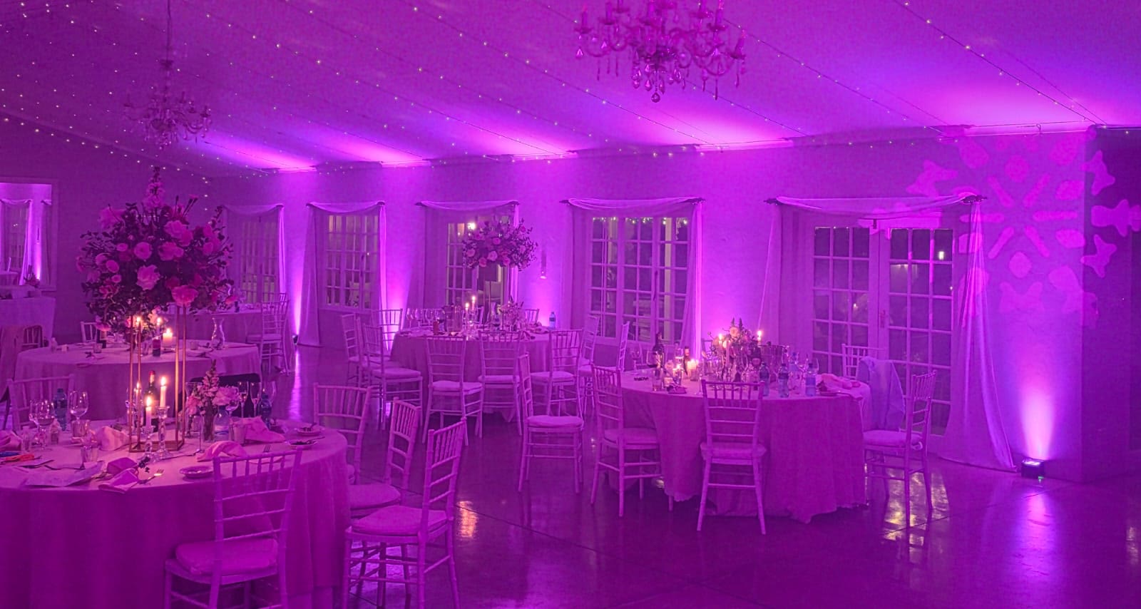 Dramatic magenta and purple uplighting throughout Providence reception hall with round tables featuring tall floral centerpieces with candles, white Tiffany chairs, crystal chandeliers, white French doors creating bold vibrant wedding atmosphere