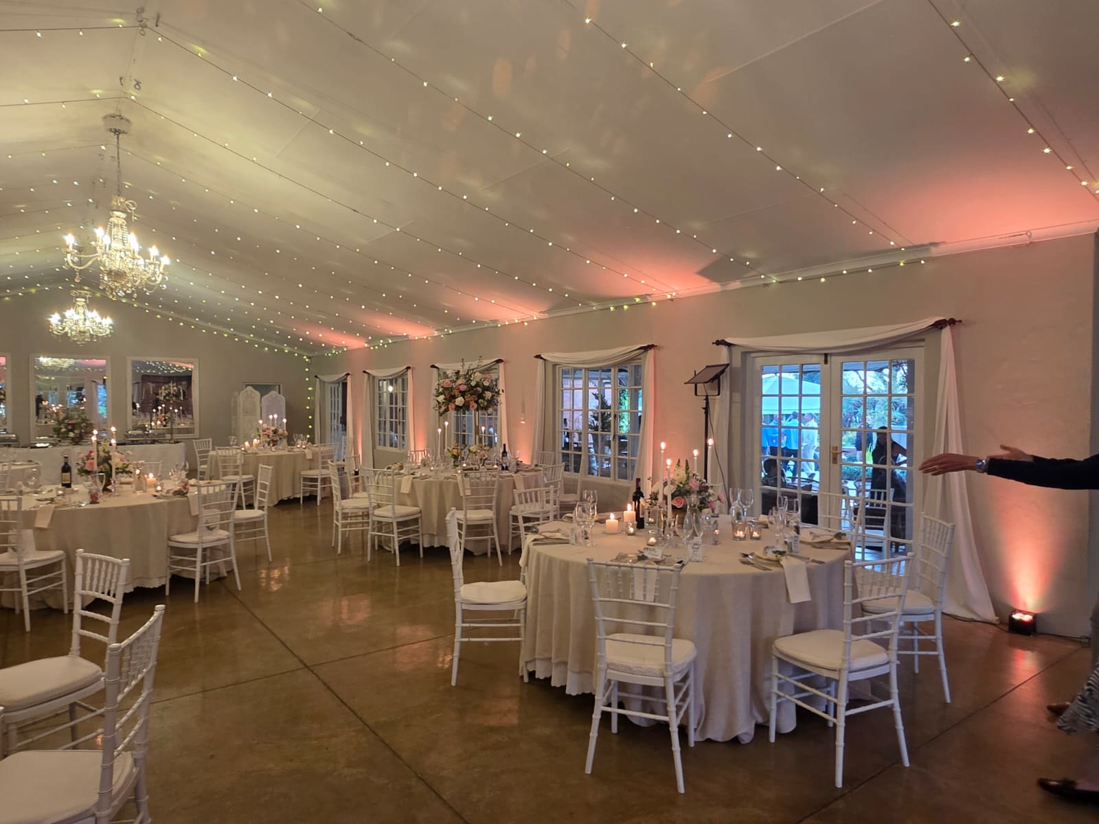 Providence reception hall set for wedding dinner with round tables, white tablecloths and Tiffany chairs, soft pink uplighting along walls, multiple crystal chandeliers, tall floral centerpieces, fairy lights creating romantic KZN Midlands atmosphere