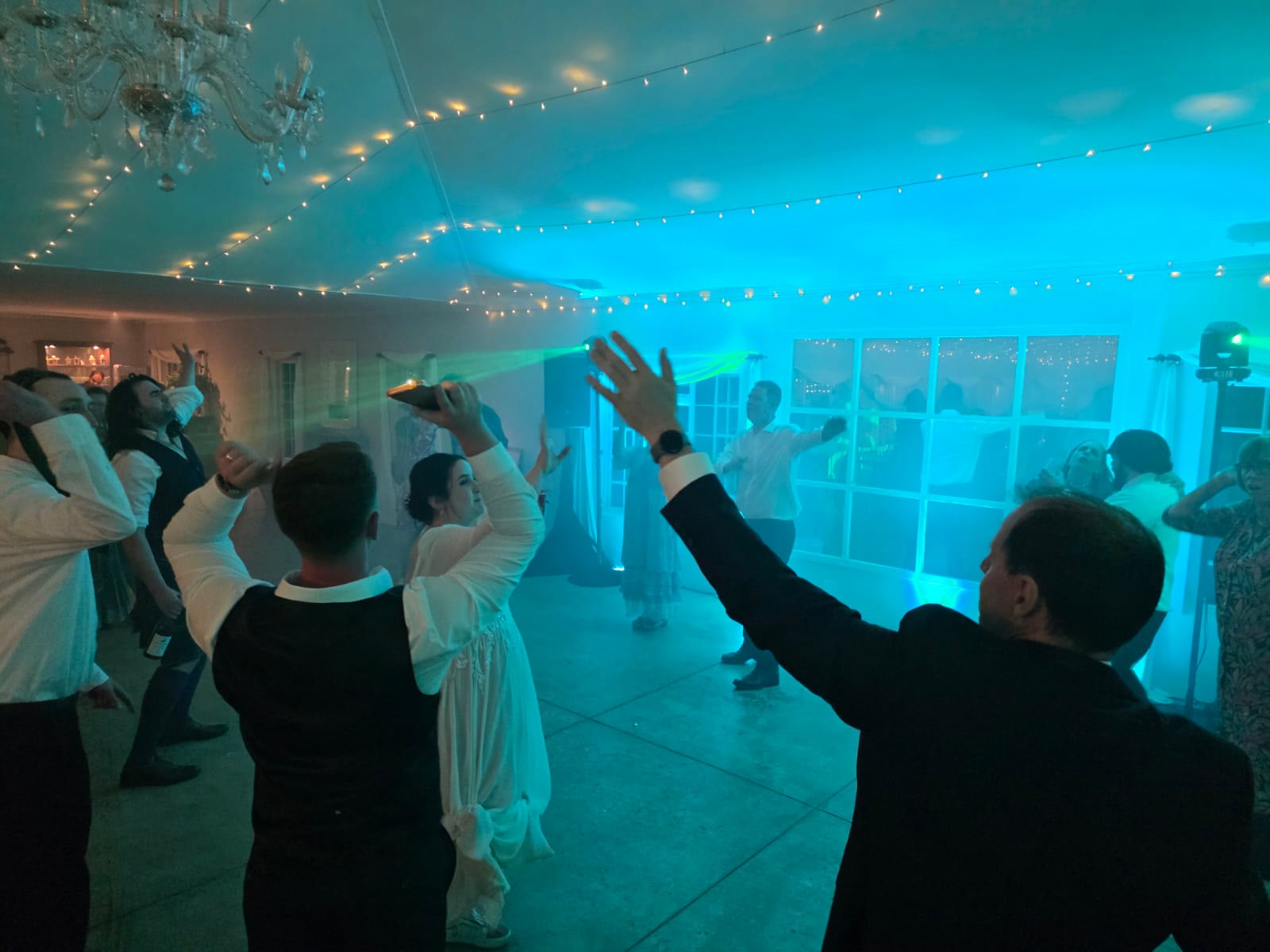 Wedding guests dancing with hands raised at Providence venue with stunning turquoise and cyan uplighting washing walls and ceiling, chandeliers overhead, white French doors, creating vibrant party atmosphere at Nottingham Road wedding